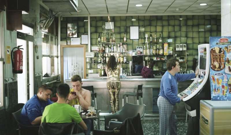 LA Kurzfilm DISCO RESPLANDECE © Chema García: Eine Bar, im Vordergrund 3 junge Männer an einem Tisch, rechts ein junger mann an einem Spielautomat, am Thresen eine junge Frau.