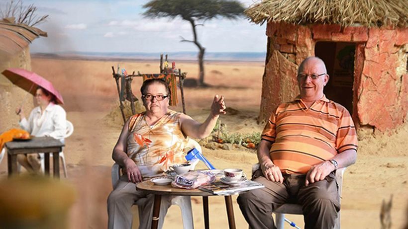 Ein Mann und eine Frau sitzen auf Gartenstühlen an einem kleinen Tisch, auf dem Tisch stehen Kaffeetassen. Im Hintergrund ist eine Wüstenlandschaft und eine Lehmhütte zu sehen.