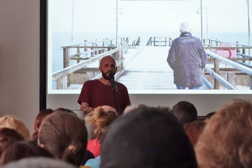 Beitragsbild für den Nachwuchstag KONTAKT der MDM: Ein junger Mann spricht vor Publikum, hinter ihm eine Leinwand, auf der ein Seebrücke zu sehen ist, darauf ein älterer Mann.