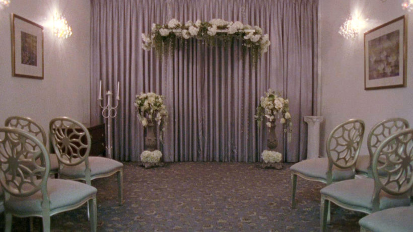 Filmstill DESERT MIRACLES: ein Blick in eine amerikanische Hochzeitskapelle, weiße Dekoration, Stühle und Blumen, zwei Bilder an den Wänden.