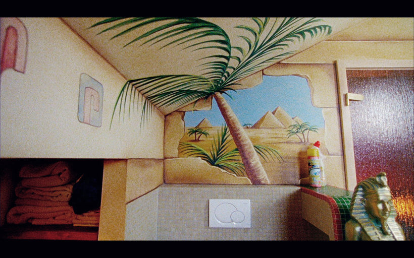 Filmstill SONNTAG BÜSCHERHÖFCHEN 2: ein Badezimmer mit ägyptischer Dekoration - ein Bild von Pyramiden, eine Palma, eine Pharao-Büste. Im Hintergrund ein WC-Reinigungsmittel.