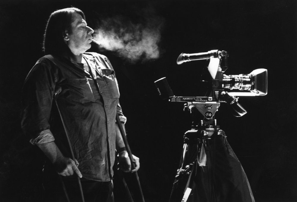 Eine schwarz-weiß-Aufnahme: ein Mann mit Gehhilfen unter beiden Armen und Zigarette im Mund steht neben einer Filmkamera.