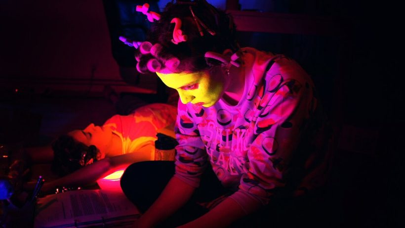 Filmstill CALL OF BEAUTY: zwei junge Frauen im Halbdunkel - eine im Vordergrund, ihr Gesicht ist mit einer gelben Gesichtsmaske bedeckt, sie trägt einen pinkfarbenen Pullover und Lockenwickler, sie schaut auf einen Computer-Monitor. Die andere Frau liegend im Hintergrund.