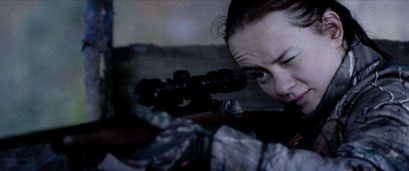 Filmstill RÅ: Ein junges Mädchen hält eine Jagdwaffe im Anschlag, sie schaut durch das Zielfernrohr. Das linke Auge ist zugekniffen.
