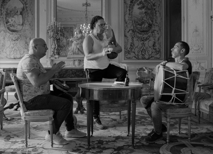 Ein Bild in schwarz-weiß. Drei Musiker in einem barocken Raum, zwei Trommler sitzend, ein Dudelsackspieler stehend an einem runden Tisch.